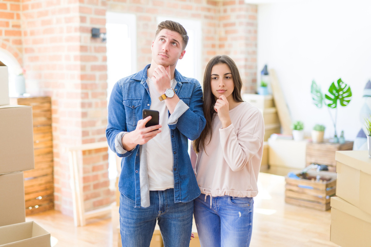 Beautiful young couple moving to a new house using smartphone serious face thinking about question, very confused idea